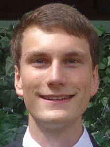 Fritzsche mit kurzem braunem Haar, freundlich lächelnd, vor einem grünen Pflanzenhintergrund.